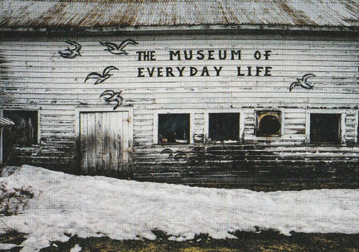 Wooden building with peeling white paint sits behind a partially snow covered ground. On the building are black painted seagulls that are flying and the hand-painted words 'The museum of every day life'.